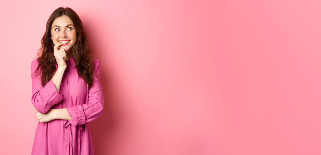 Coquettish young woman looking thoughtful aside, smiling and biting finger while thinking, imaging something, standing over pink background Nachdenkliche Frau; über Krankenversicherun