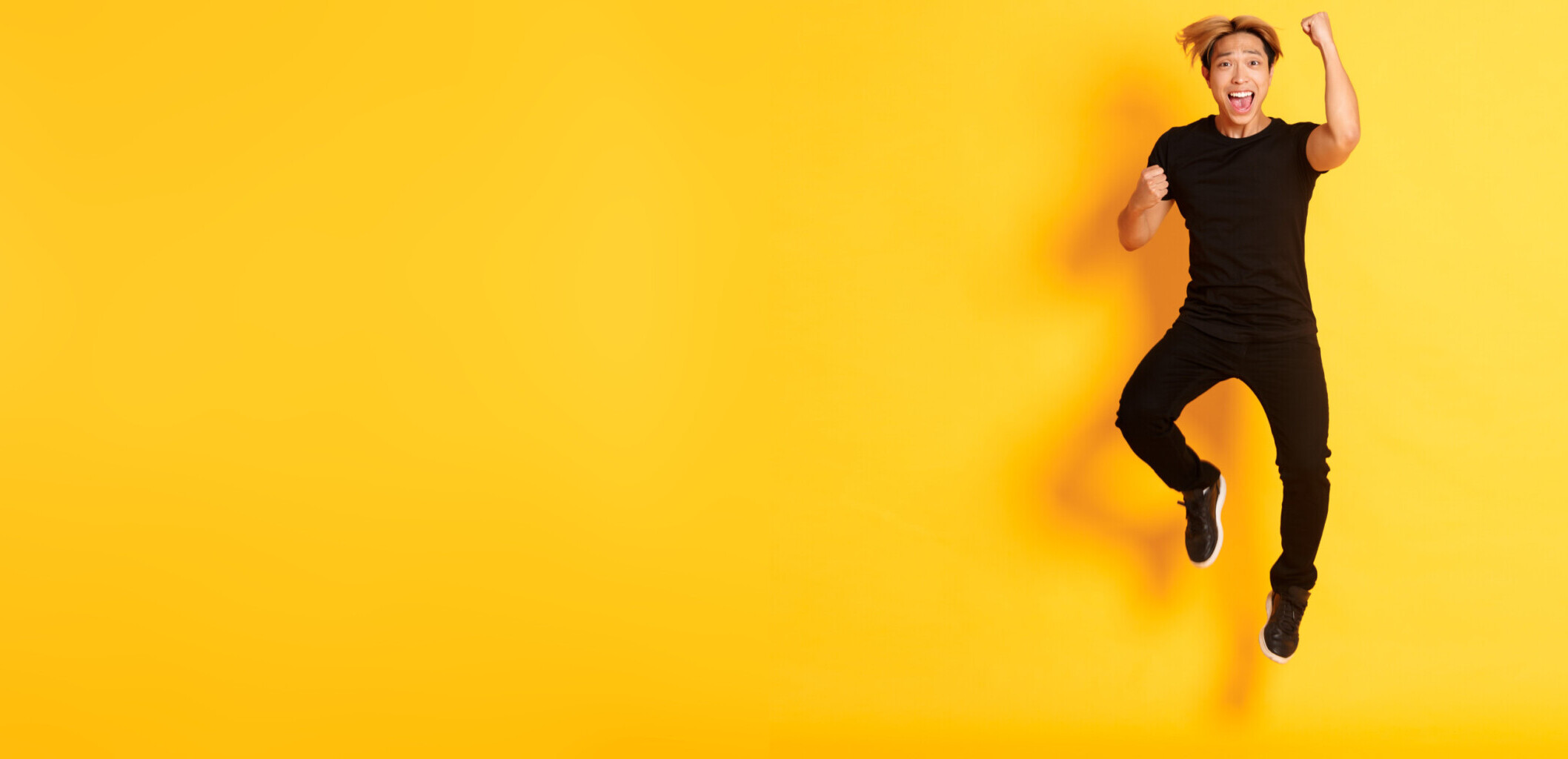 Full-length of happy attractive asian guy in black clothes jumping and celebrating victory, achieve goal, standing yellow background