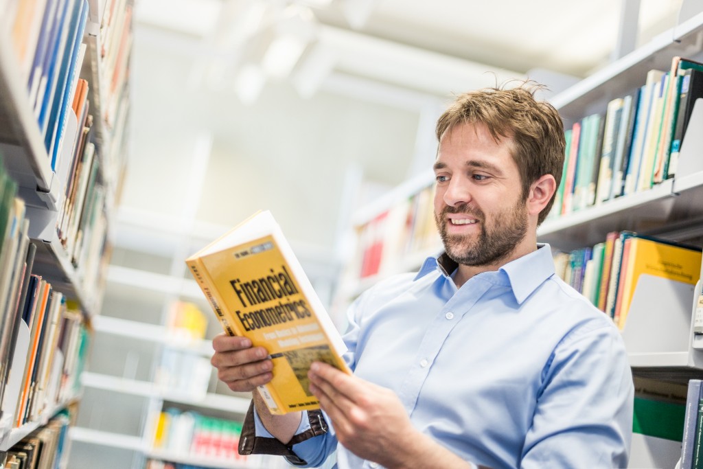 M. S. Ein Mann steht in einer Bibliothek und liest ein Finanzbuch.