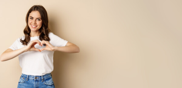 Lovely woman showing heart, love sign and smiling, standing over beige background in white t-shirt, beige background Junge Frau formt mit den Händen ein Herz und lächelt.